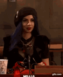 a woman with blue hair and red gloves is sitting in front of a microphone with the name nelli g below her