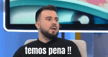 a man with a beard is sitting in front of a television and says temos pena !