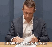 a man in a suit is sitting at a table writing on a piece of paper with the words coming in as a z card .