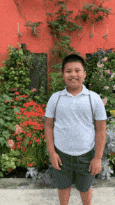 a boy wearing a white shirt and green shorts stands in front of a wall of flowers