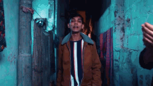 a young man in a brown jacket and striped shirt is standing in a dark alleyway .