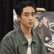 a young man wearing a denim jacket and a white shirt is standing in front of a poster of a group of men .