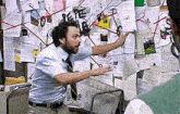 a man in a blue shirt and tie is pointing at a bulletin board with a lot of papers on it .