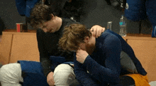 two men are sitting next to each other in a locker room and one of them is holding the other 's head .