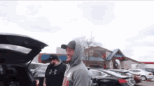 a man in a hooded sweatshirt is standing next to a car with the trunk open .