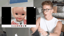 a man wearing glasses looks at a picture of a doll on a laptop screen