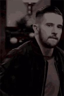 a man with a beard is wearing a black jacket and a gray shirt and looking at the camera .