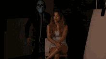 a woman is sitting in a chair while a man wearing a blue mask stands behind her