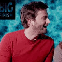 a man wearing a red shirt is laughing in front of a sign that says big finish
