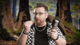 a man wearing glasses and a checkered shirt is holding a microphone in front of trees