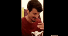a man with a beard is wearing a red shirt and holding a piece of food in his hand .