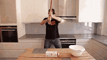 a man in a black shirt is holding a wooden bat in a kitchen .