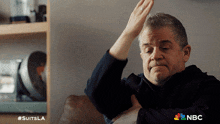 a man is sitting on a couch with his hands in the air and a nbc logo in the corner