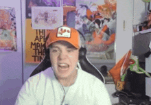 a man wearing an orange hat is sitting in front of a wall with a poster that says " the art may make the tin " on it