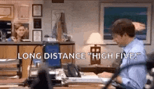 a man is sitting at a desk in an office with a laptop and a woman is behind him .