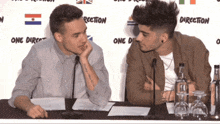 two men sit at a table in front of a one direction sign