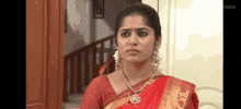 a woman wearing a red saree and a gold necklace is standing in front of a staircase .