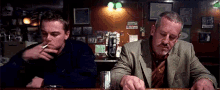 a man is smoking a cigarette while sitting at a bar next to another man .