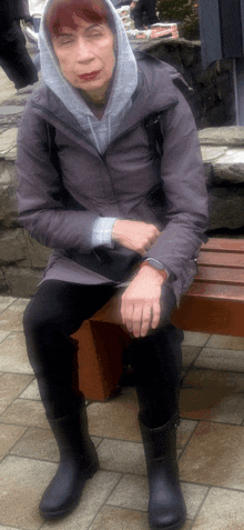 a woman wearing a hooded jacket and black boots sits on a wooden bench