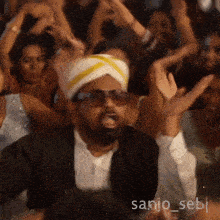 a man wearing sunglasses and a turban stands in front of a crowd with the name sanjo_sebi written on the bottom