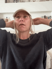 a woman wearing a baseball cap and a black sweater has her hands on her head