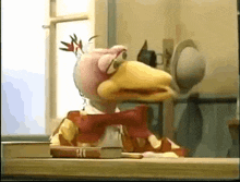 a chicken puppet is sitting at a table with books and a hat .