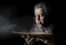 a man wearing glasses is smoking a cigarette while reading a book in a dark room .