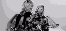 a man wearing sunglasses is sitting on the back of a car next to another man wearing a camouflage jacket .