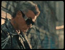 a man wearing sunglasses and a leather jacket stands in front of a graffiti wall