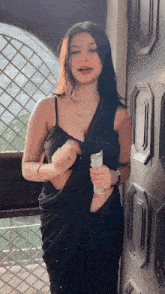 a woman in a black saree is standing in front of a window and holding a bottle .