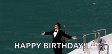 a man in a tuxedo and bow tie is standing on a boat with his arms outstretched .