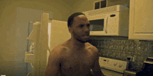 a shirtless man stands in a kitchen with a microwave and refrigerator