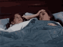 two women are laying in bed with a blue blanket . one of the women is touching the other 's forehead .