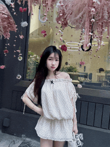 a woman wearing a white off the shoulder top stands in front of a window with pink flowers hanging from it