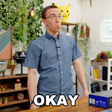 a man wearing glasses and a blue shirt is standing in front of a table with the word okay on it