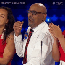 a man wearing glasses and a tie stands in front of a microphone with a #familyfeud canada hashtag on the bottom