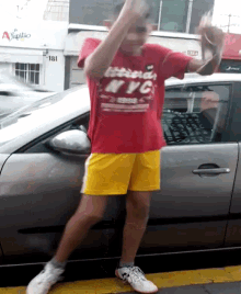 a man wearing a red nyc shirt and yellow shorts is standing in front of a car