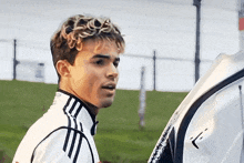 a young man wearing a white adidas jacket stands in a field
