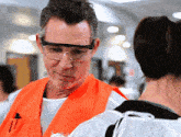 a man wearing safety glasses and an orange vest with a pen in his pocket