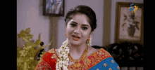 a woman in a blue and red saree is standing in a living room wearing a garland of flowers .