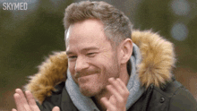 a man wearing a fur hooded parka is clapping his hands .