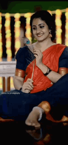 a woman in a red and blue saree is holding a peacock feather in her hand .