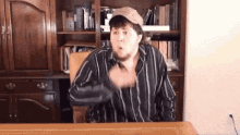 a man wearing a hat and a striped shirt is sitting at a desk in front of a bookshelf .