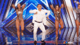 a man in a white suit is dancing on a stage with two women in silver leotards behind him .