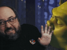 a man wearing glasses and a black shirt with a smiley face on it is standing next to a shrek statue