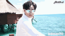 a man wearing sunglasses and a white sweater is standing in front of a body of water