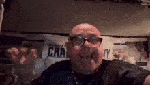a bald man wearing glasses is making a funny face while sitting in front of a wall .