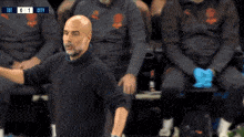 a bald man in a black sweater stands in front of a group of men wearing grey jackets and gloves