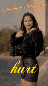 a woman in a black jacket and skirt is standing in front of a body of water and the name kurt is on the bottom