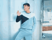 a young man in a blue and black sweater is waving his hand while leaning against a wall .